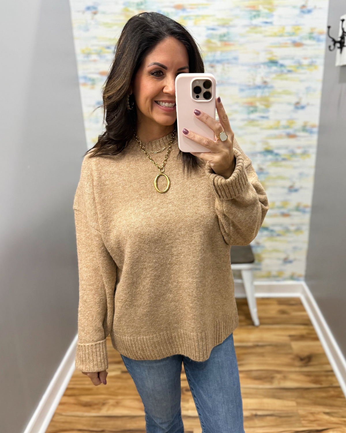A woman takes a mirror selfie wearing a beige knit crewneck sweater and blue jeans in a home setting with wood floors and a patterned wallpaper background.