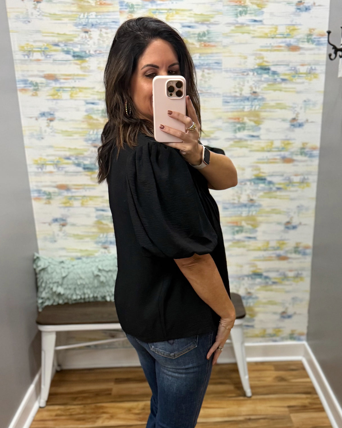 A woman takes a mirror selfie wearing a black puff-sleeve blouse and blue jeans in a fitting room with patterned wallpaper and a bench.