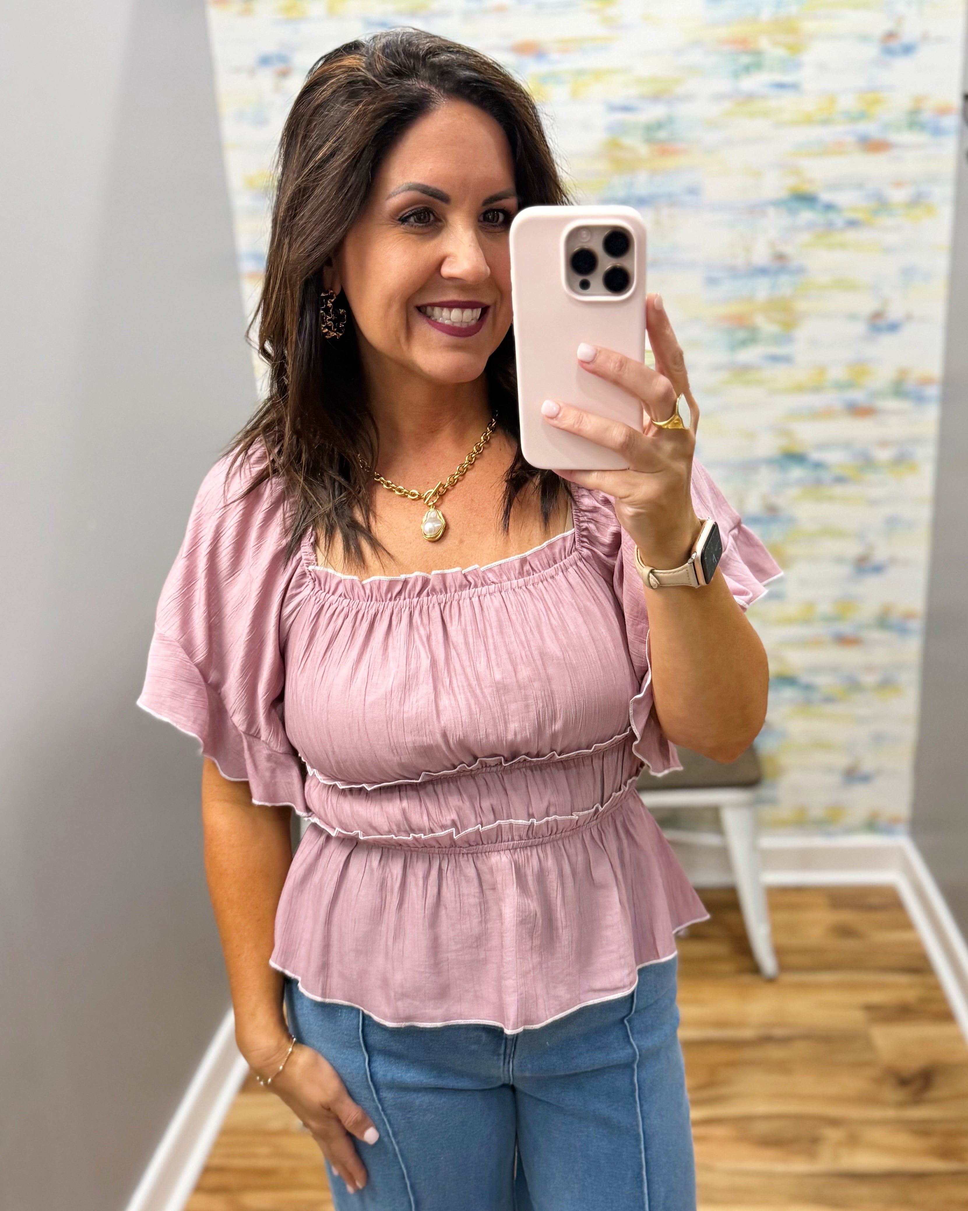 Woman taking a mirror selfie wearing a pink top and blue jeans, with a colorful wall in the background.
