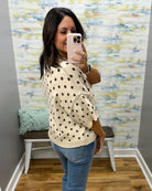 A woman takes a mirror selfie in a fitting room wearing a cream knit sweater with brown polka dots and blue jeans.