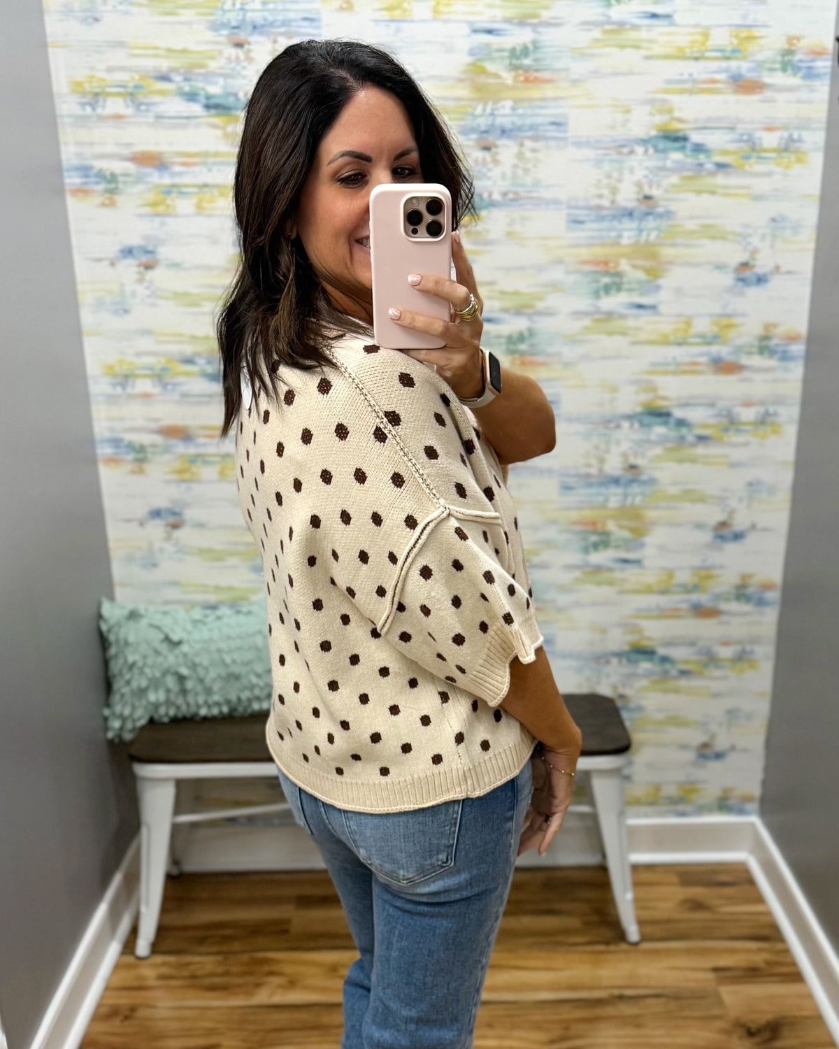 A woman takes a mirror selfie in a fitting room wearing a cream knit sweater with brown polka dots and blue jeans.