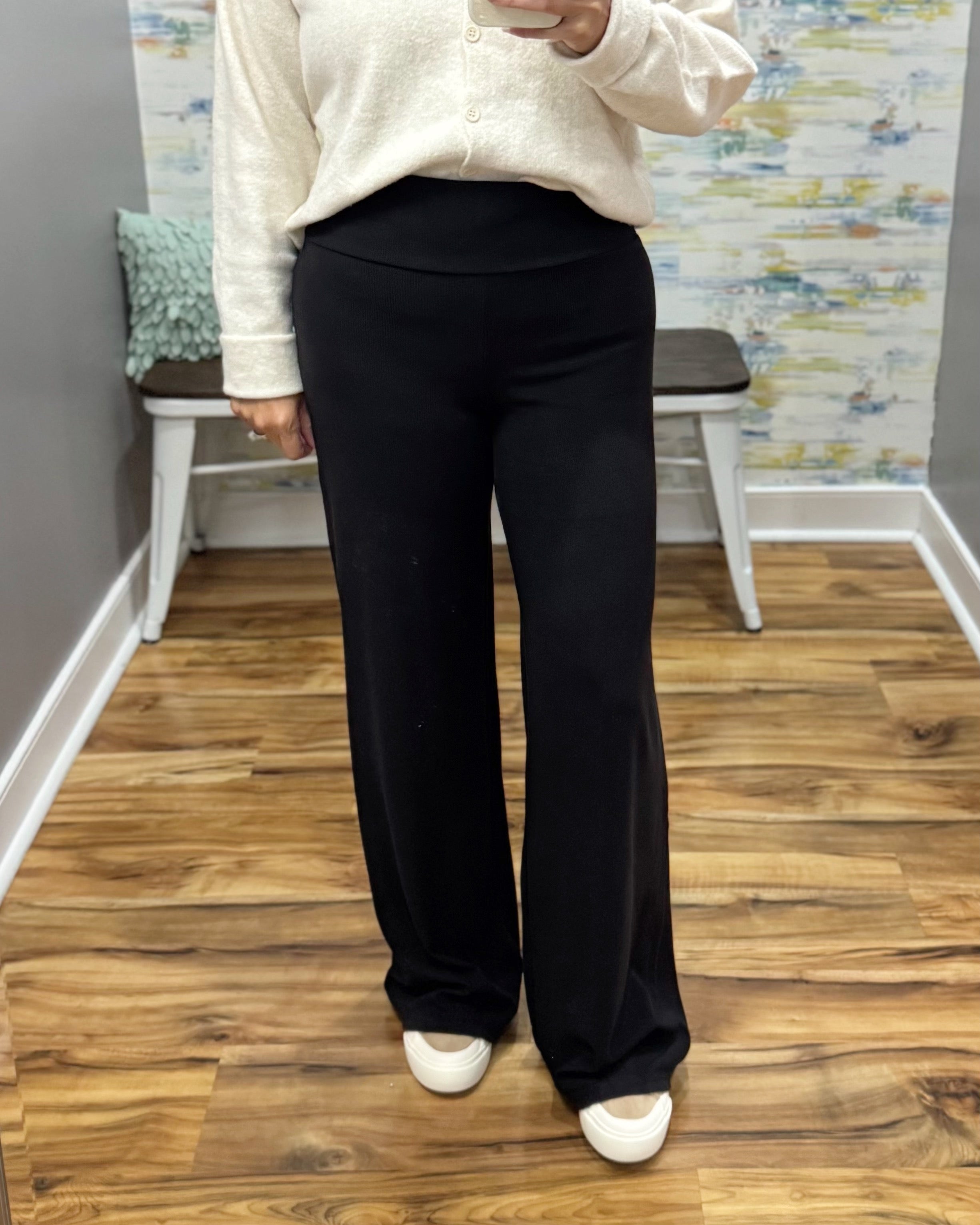A person wearing black wide-leg pants, a cream button-front top, and white sneakers, standing in a room with wood floors and a bench in the background.
