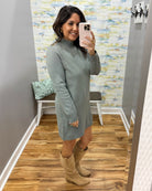 A woman takes a mirror selfie in a fitting room wearing a sage green mock-neck knit sweater dress and beige cowboy boots.
