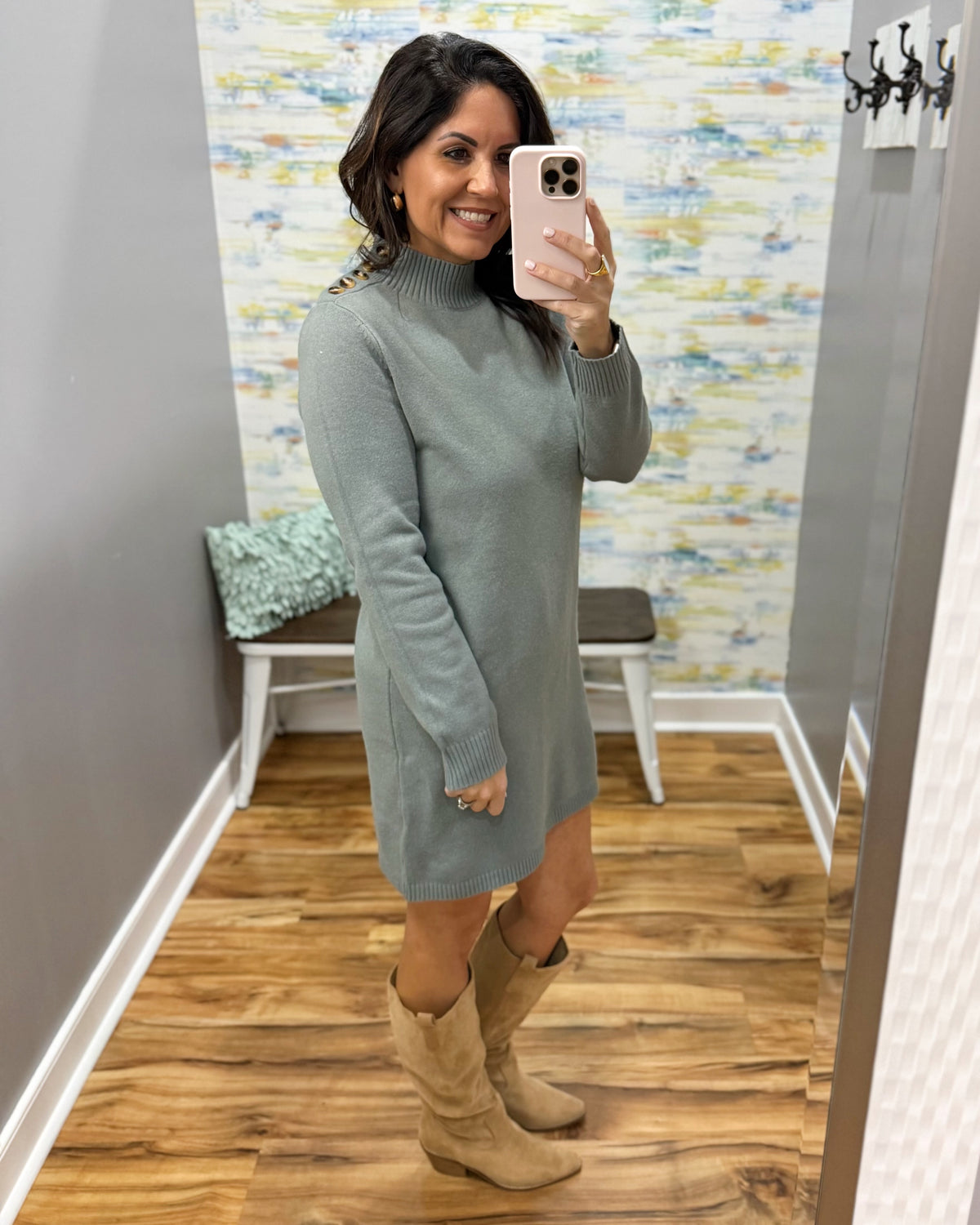 A woman takes a mirror selfie in a fitting room wearing a sage green mock-neck knit sweater dress and beige cowboy boots.