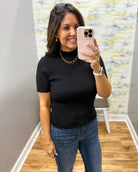 A woman taking a mirror selfie in a boutique, wearing a black short-sleeve ribbed top with a scalloped mock neck and blue jeans.