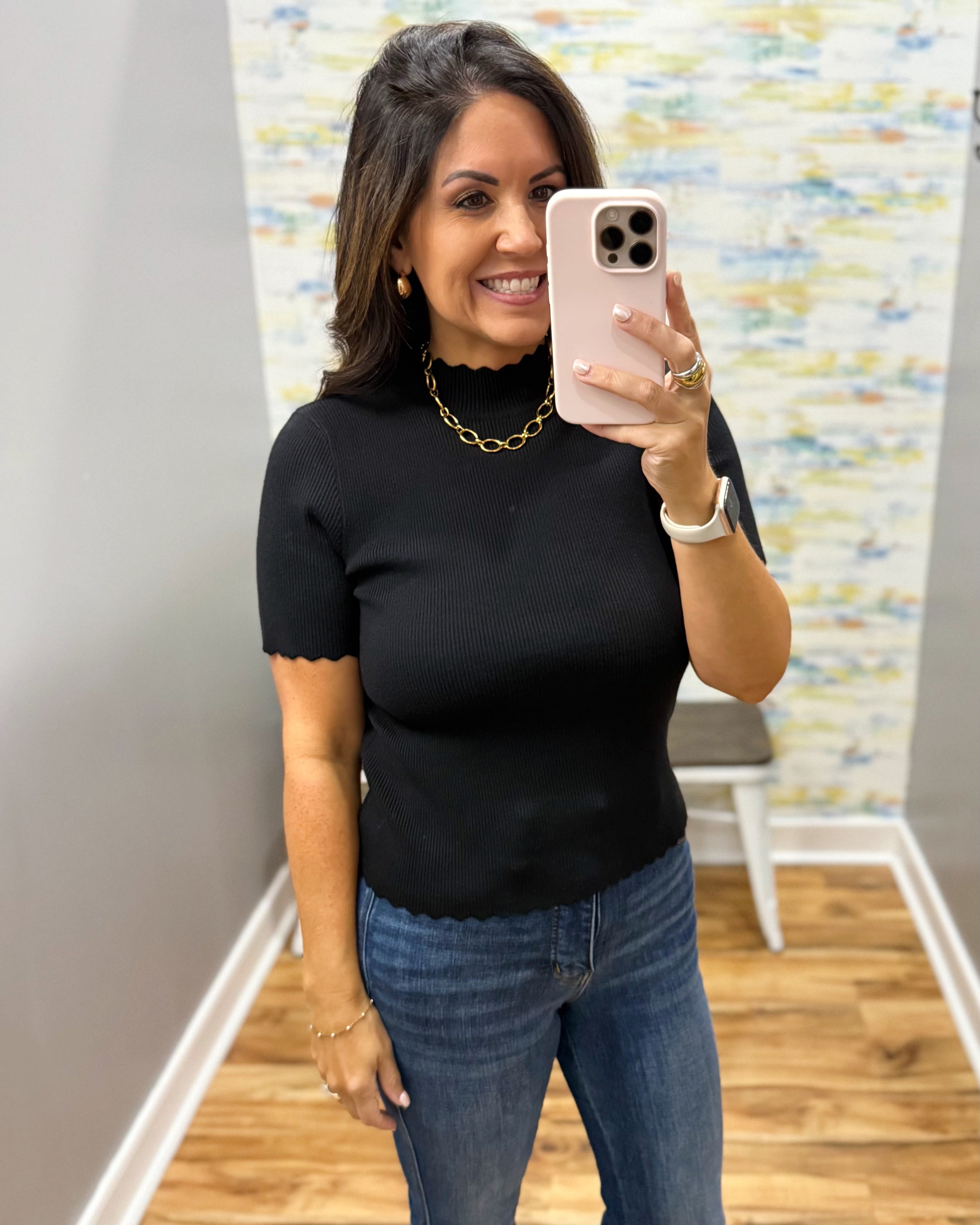 A woman taking a mirror selfie in a boutique, wearing a black short-sleeve ribbed top with a scalloped mock neck and blue jeans.