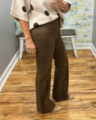 A person wearing brown wide-leg pants and a cream knit sweater with black polka dots, standing on a wood floor in an indoor room with a bench and abstract wall art in the background.