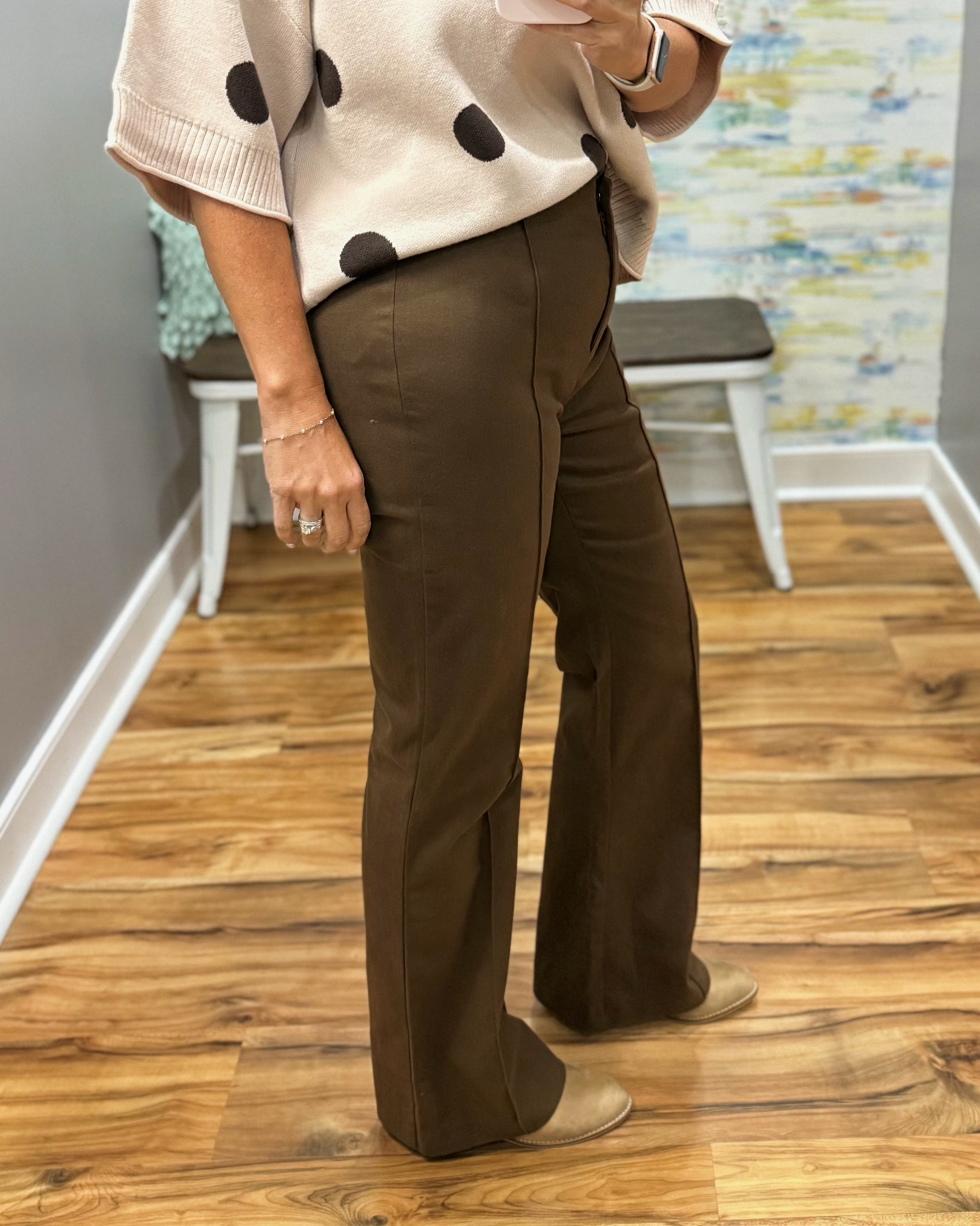 A person wearing brown wide-leg pants and a cream knit sweater with black polka dots, standing on a wood floor in an indoor room with a bench and abstract wall art in the background.