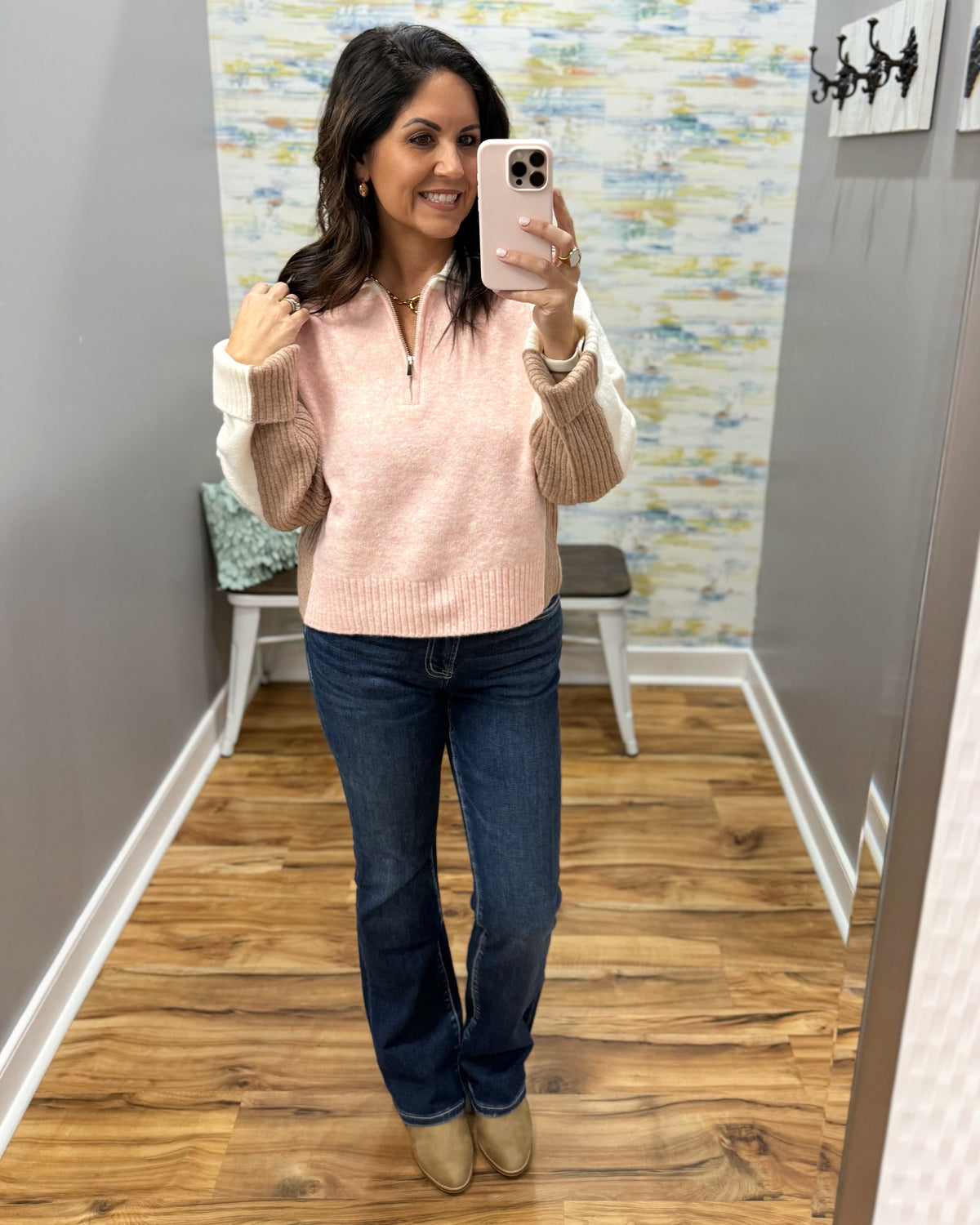 A woman takes a mirror selfie wearing a light pink ribbed quarter-zip sweater with beige knit sleeves, paired with dark blue jeans and tan ankle boots, in a home fitting room.