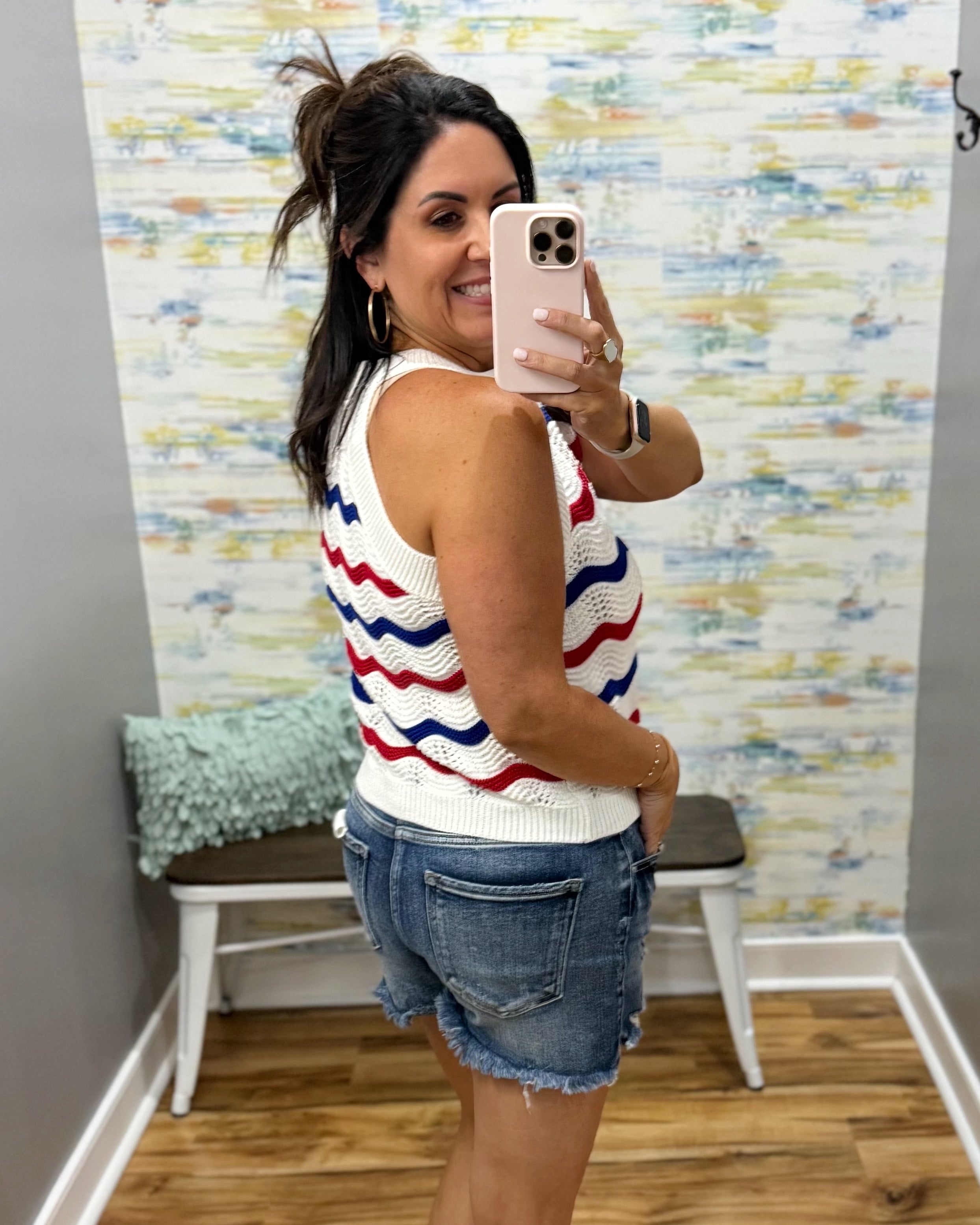 Woman taking a mirror selfie wearing a striped sleeveless top and denim shorts in a room with colorful wall art.