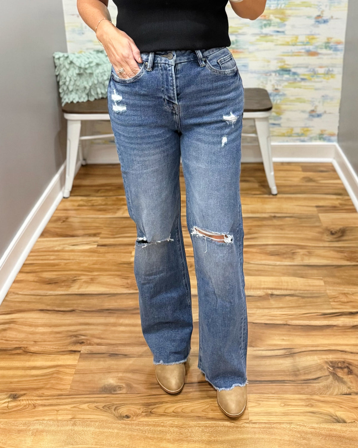 A person wearing mica wide leg distressed jeans with ripped details on the thighs and knees, paired with a black top and tan boots, standing in an indoor room with wood floors and a bench.