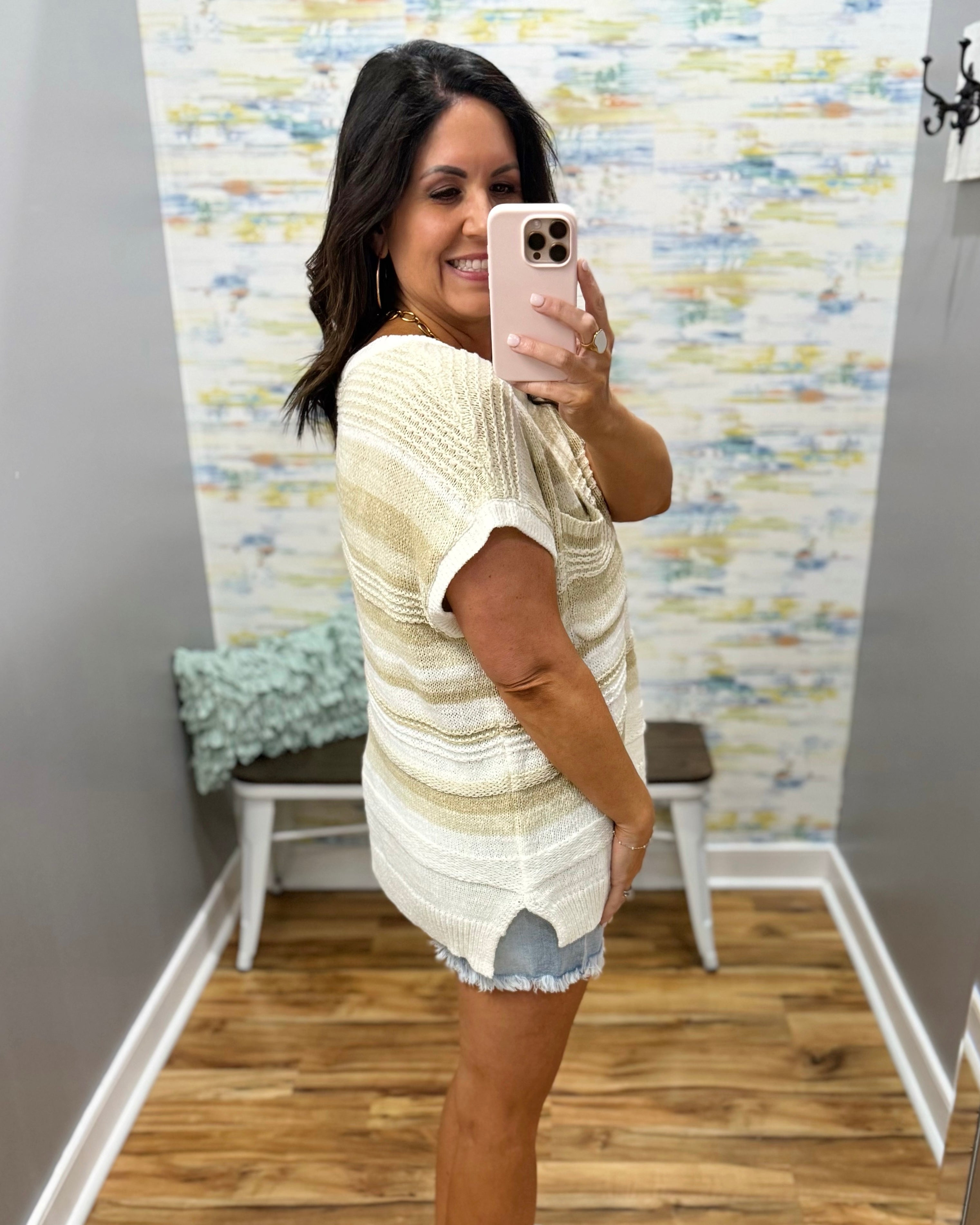 Woman taking a mirror selfie wearing a light-colored outfit with a patterned wall in the background.