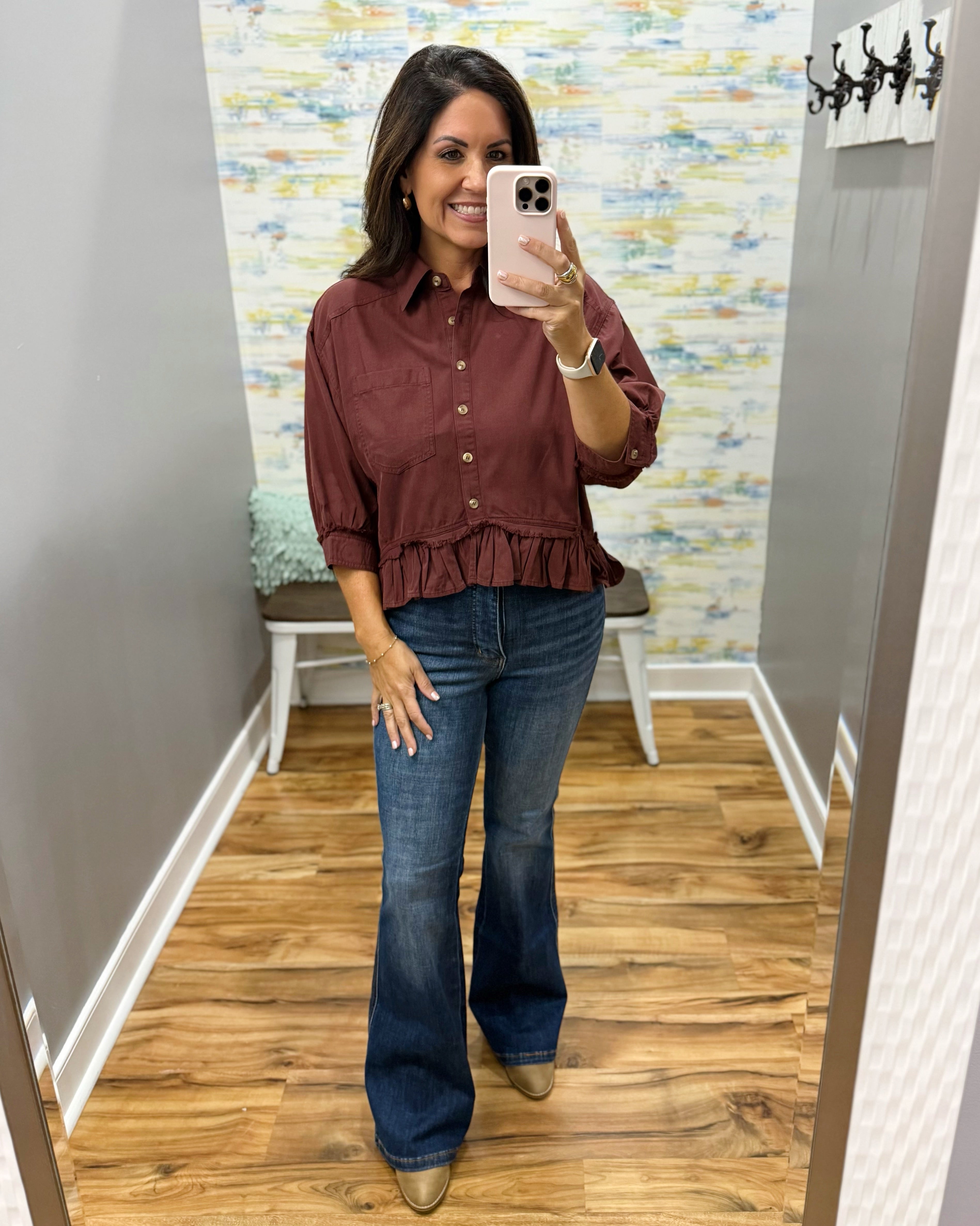 A woman takes a mirror selfie in a fitting room wearing a rust-brown ruffled button-up blouse and dark blue flare jeans.