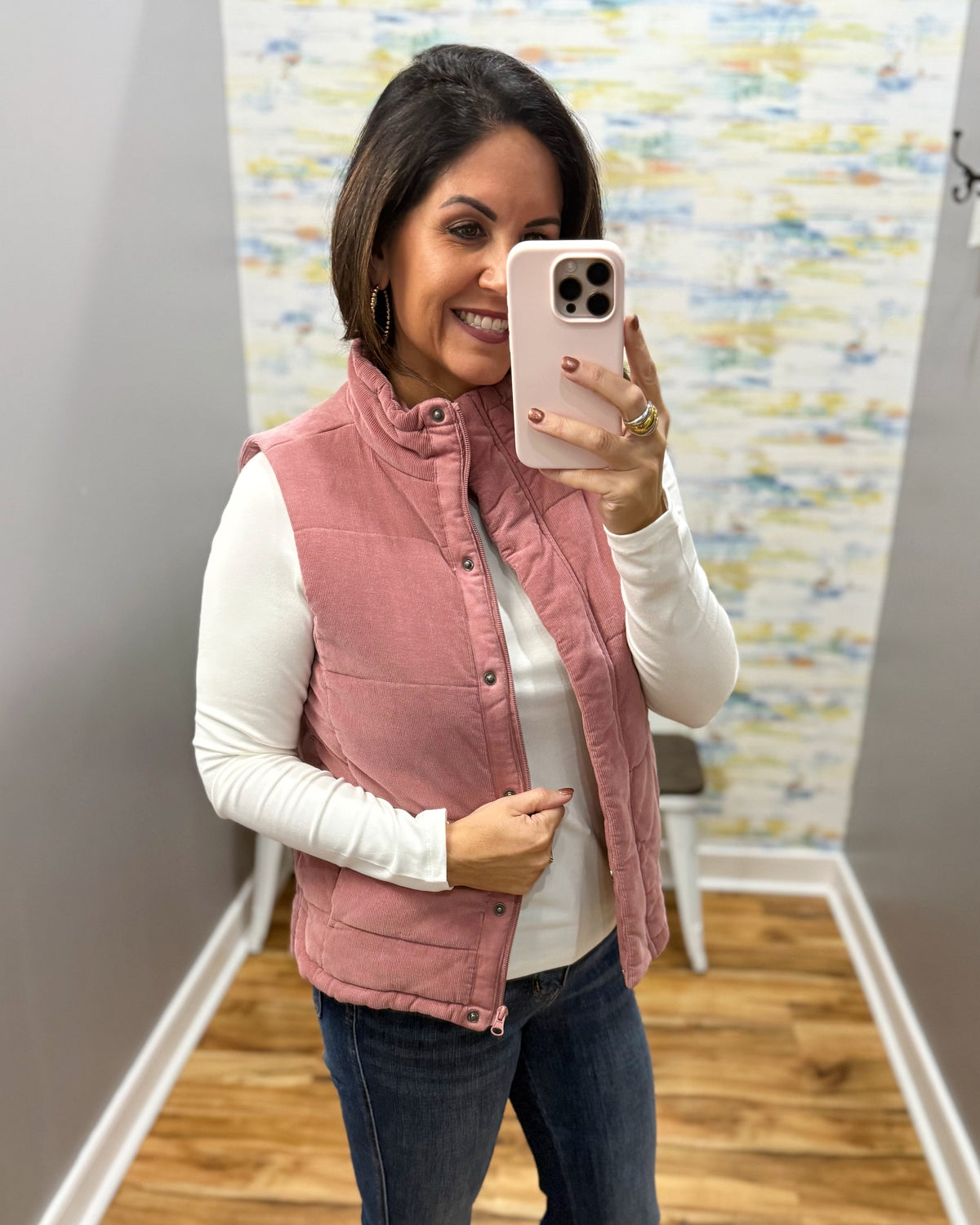 A woman takes a mirror selfie wearing a dusty rose pink quilted sleeveless vest over a white long-sleeve shirt and blue jeans.