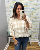 A woman taking a mirror selfie in a boutique wearing a beige and white checkered peplum blouse with three-quarter sleeves and dark blue jeans.