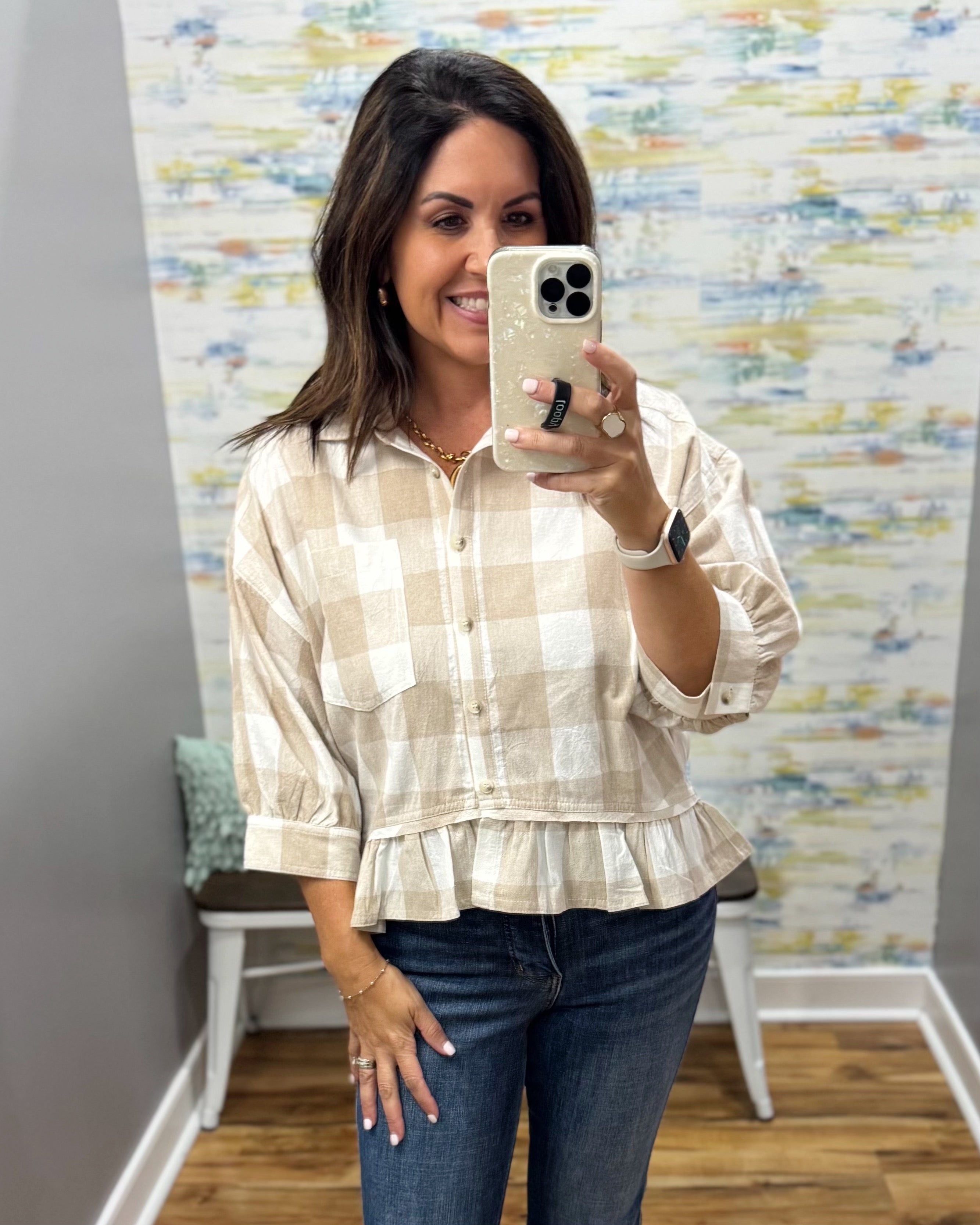 A woman taking a mirror selfie in a boutique wearing a beige and white checkered peplum blouse with three-quarter sleeves and dark blue jeans.