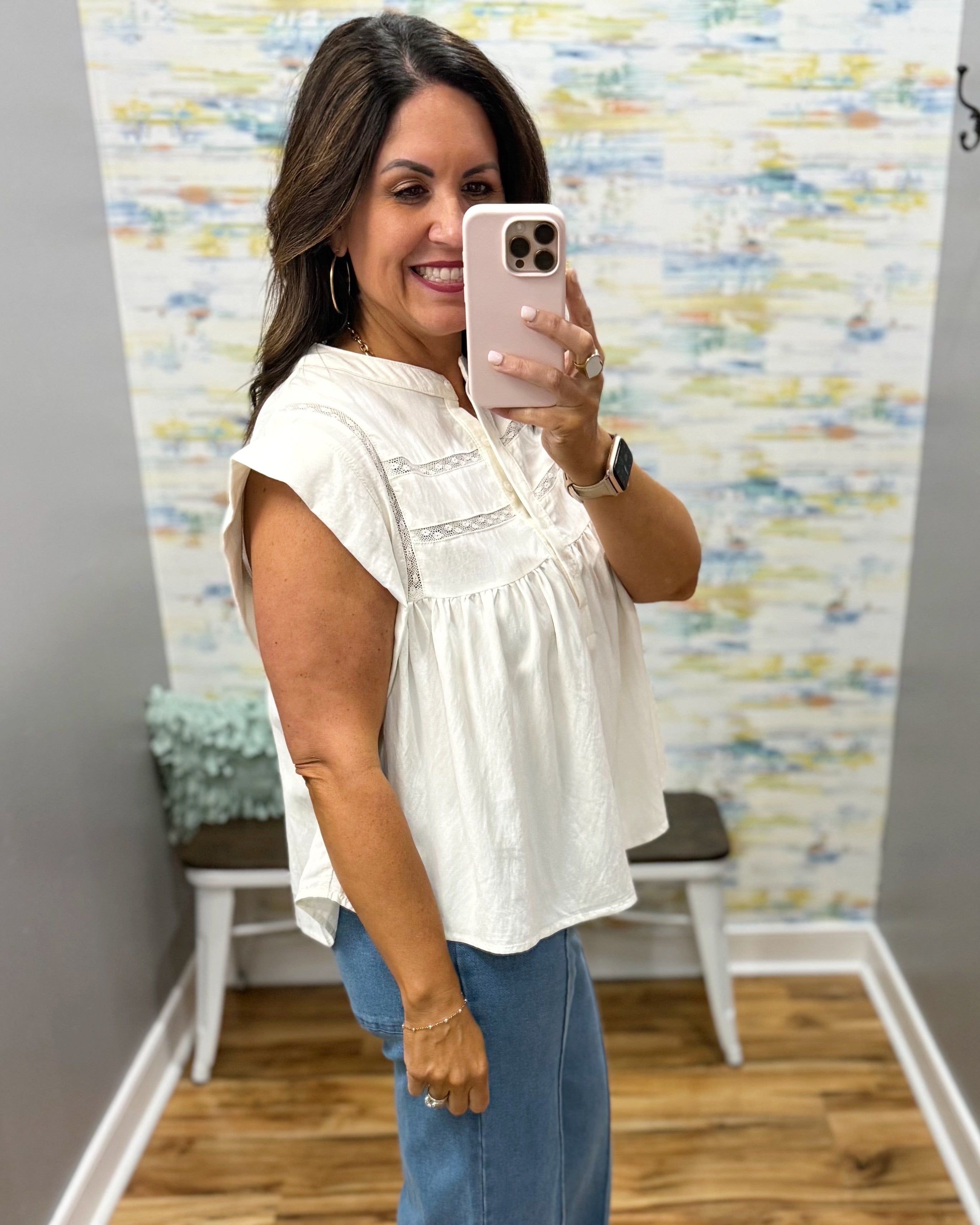 Woman taking a mirror selfie wearing a white blouse and blue jeans in a room with a colorful wall.