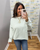 A woman takes a mirror selfie wearing a light green and white striped long-sleeve top with blue jeans, standing in a fitting room or home interior with a patterned wall and wood floor.