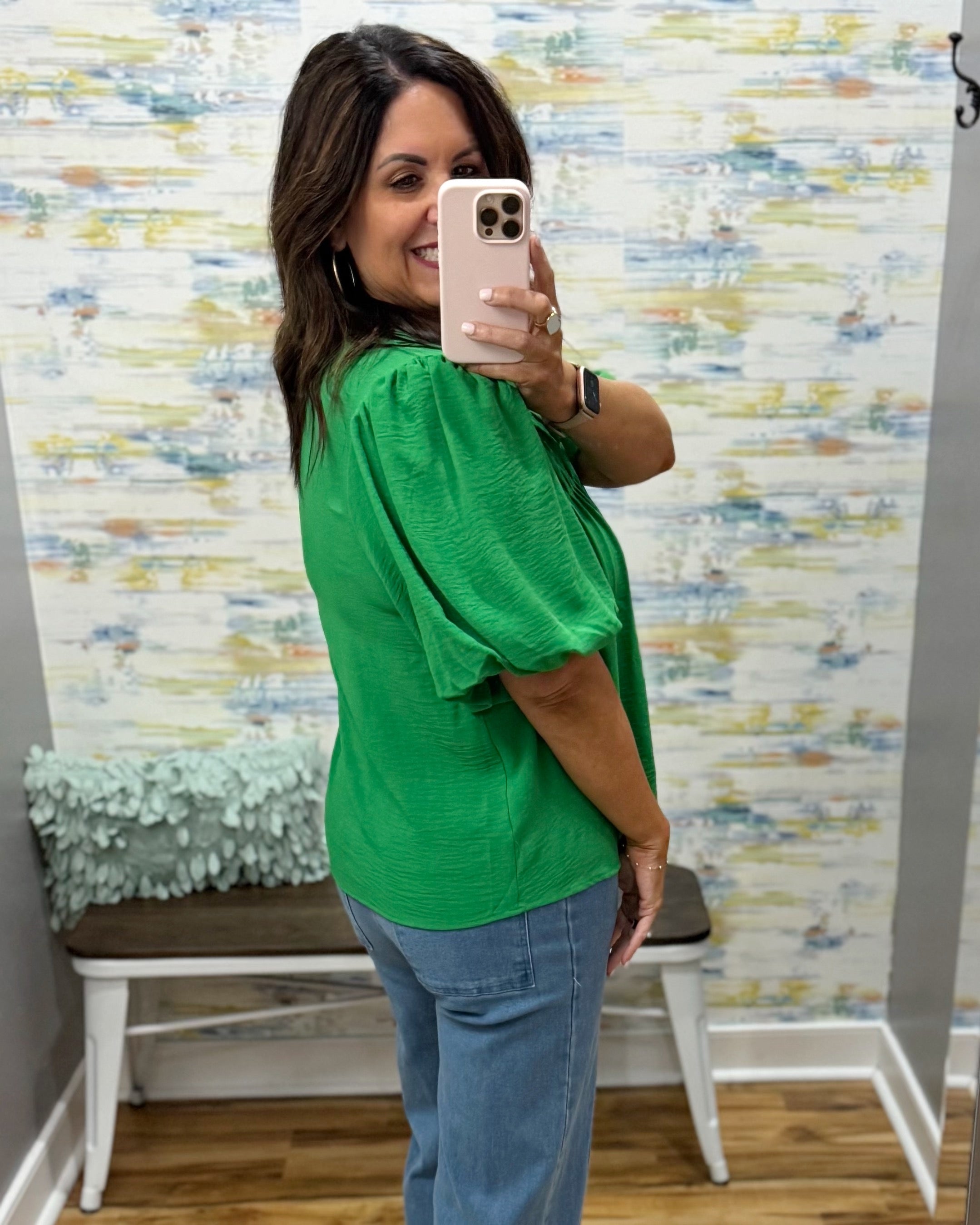 Woman taking a mirror selfie wearing a green top and blue jeans in front of a colorful wall.