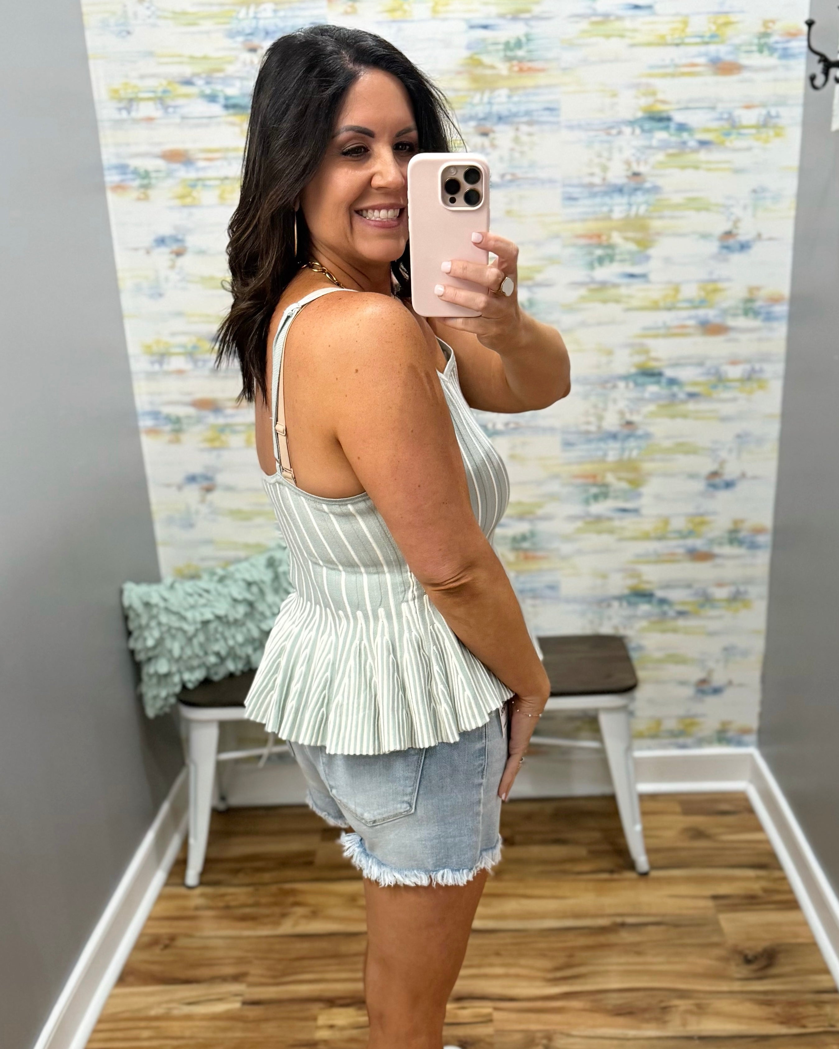 Woman taking a mirror selfie wearing a white pleated top and denim shorts in a room with patterned wallpaper.