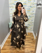 A woman takes a mirror selfie in a boutique fitting room wearing a dark brown long-sleeve maxi dress with a beige floral pattern and a matching waist tie belt.