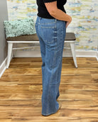 A person wearing mica wide-leg jeans, a black top, and tan pointed-toe heels, standing on a wood floor in an indoor room with a bench and abstract wall art in the background.