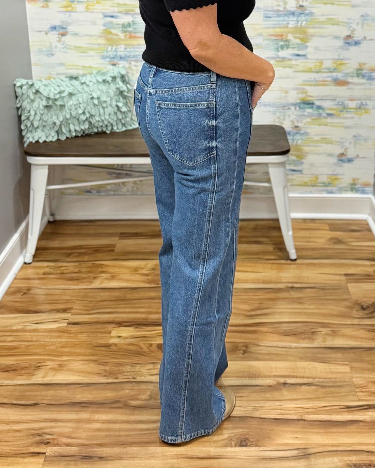 A person wearing mica wide-leg jeans, a black top, and tan pointed-toe heels, standing on a wood floor in an indoor room with a bench and abstract wall art in the background.