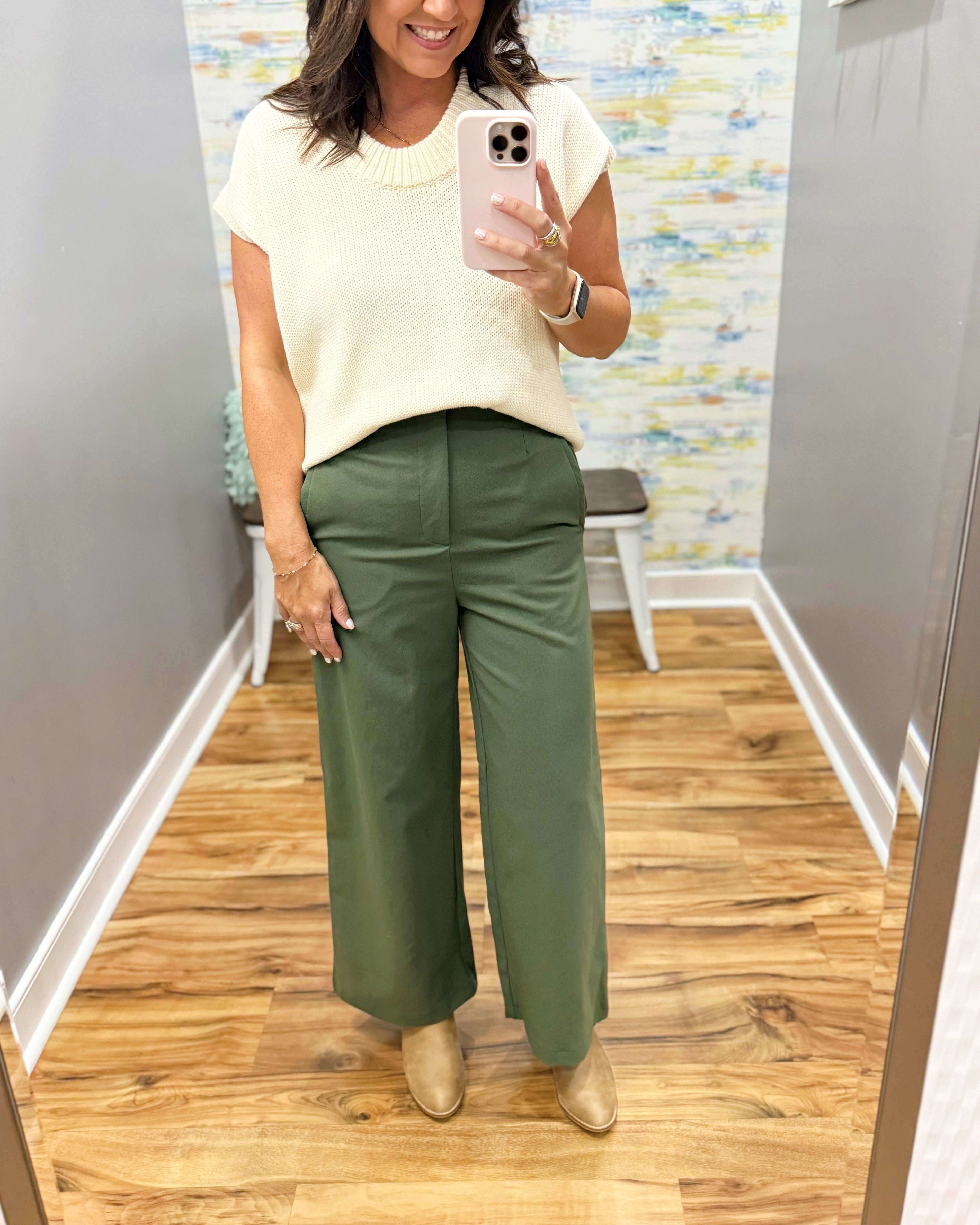 A woman takes a mirror selfie in a fitting room wearing a cream short-sleeve knit top and olive green wide-leg pants with beige ankle boots.