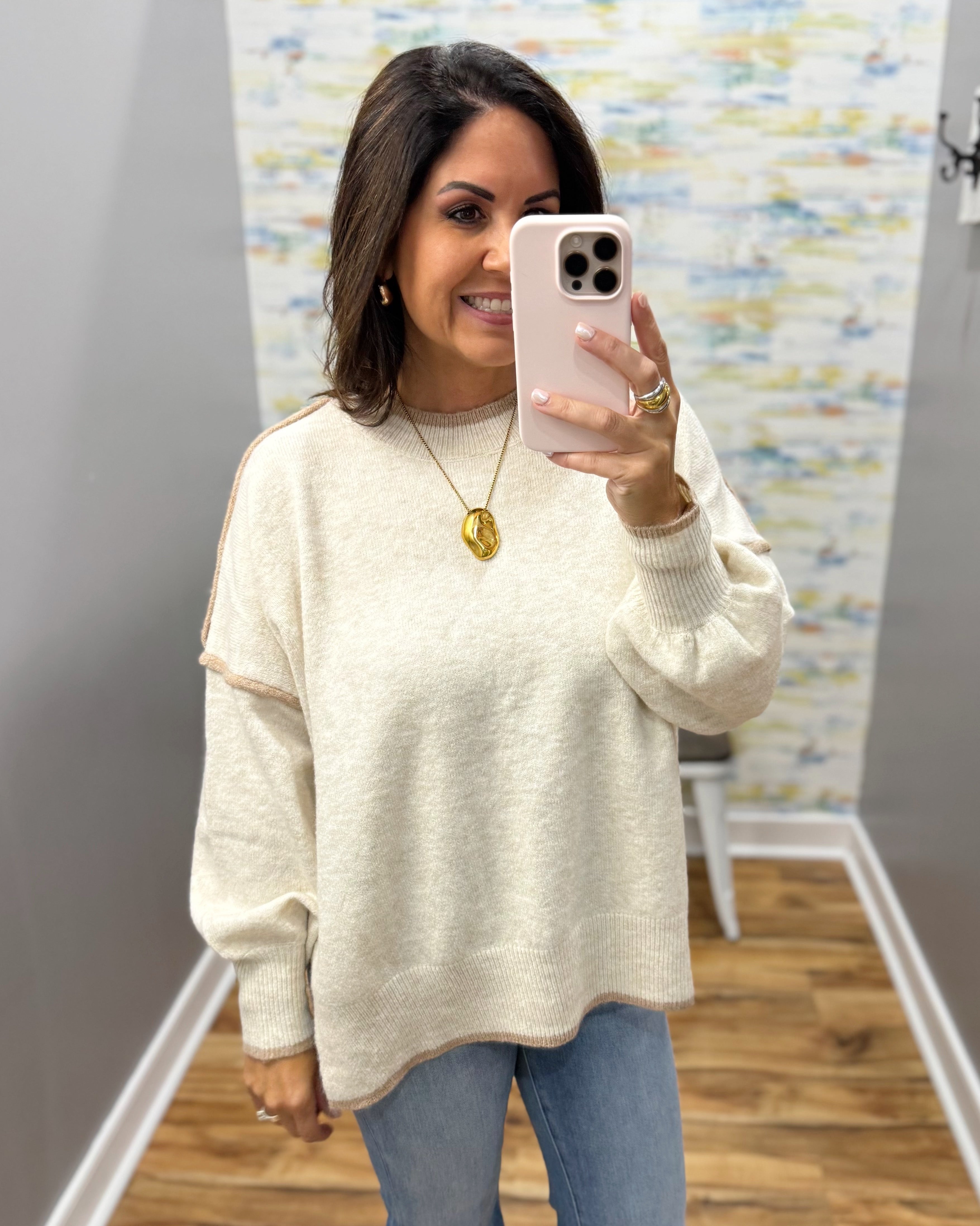 A woman takes a mirror selfie wearing a cream-colored knit sweater with beige contrast stitching along the shoulders and sleeves, paired with blue jeans.
