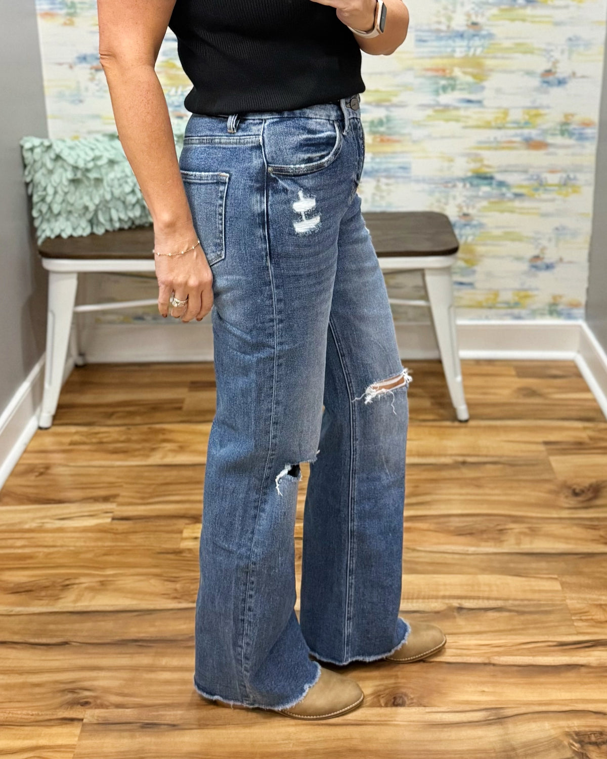A person wearing mica wide leg distressed jeans with ripped details on the thighs and knees, paired with a black top and tan boots, standing in an indoor room with wood floors and a bench.