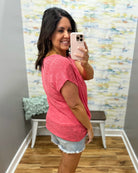 Woman taking a mirror selfie wearing a pink shirt and denim shorts in a room with colorful wall art.