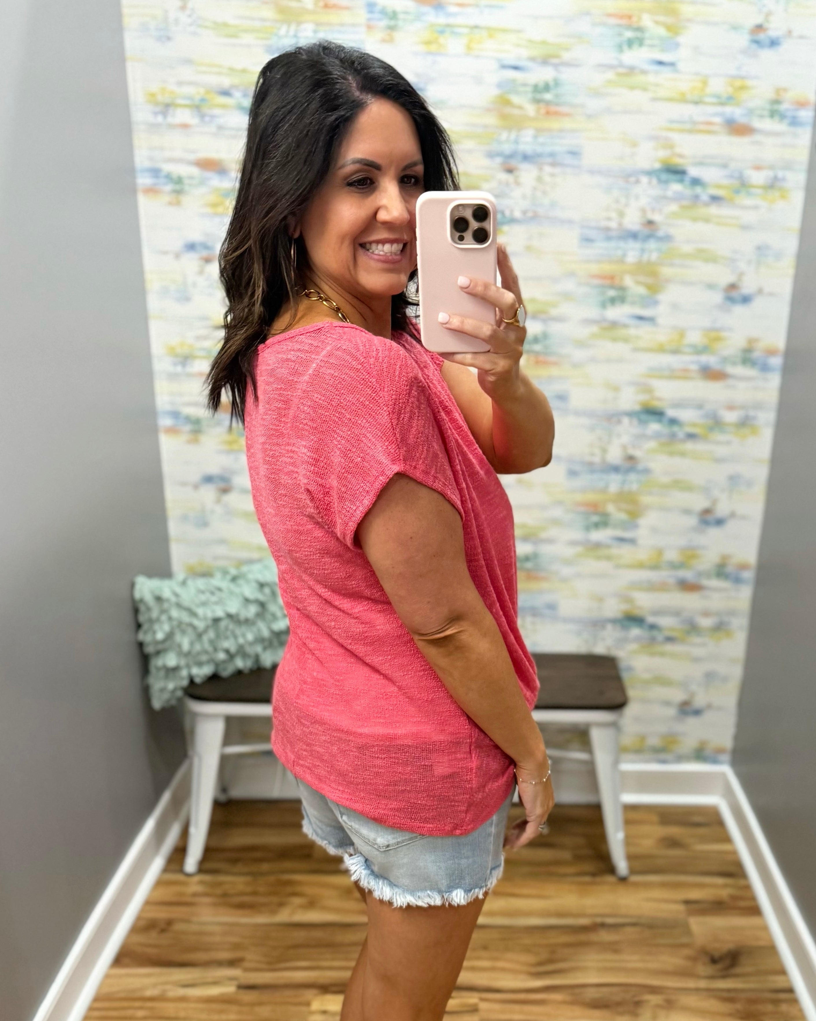 Woman taking a mirror selfie wearing a pink shirt and denim shorts in a room with colorful wall art.