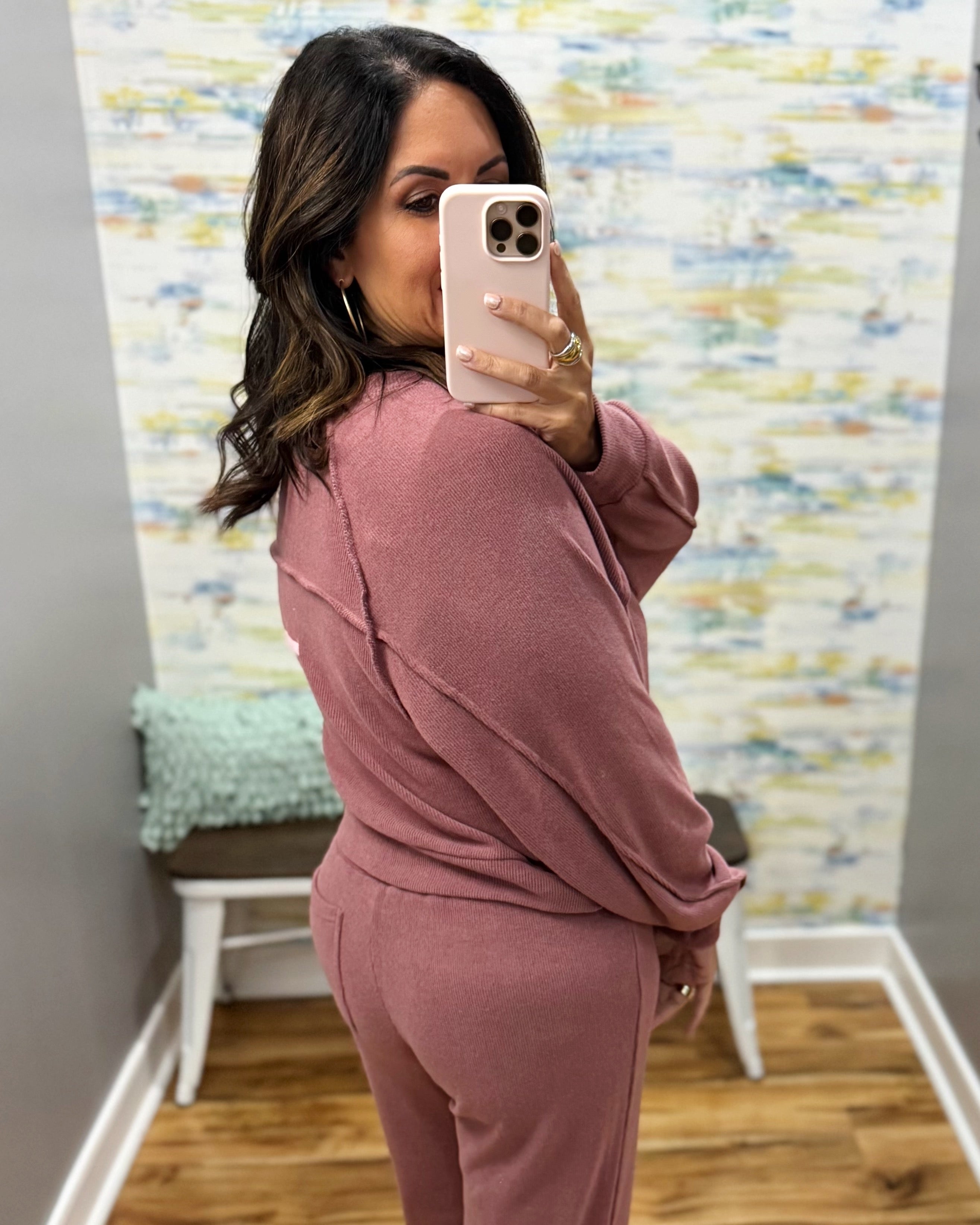 A woman takes a mirror selfie wearing a dusty rose two-piece lounge set consisting of a long-sleeve top and matching pants. The setting is a fitting room or home interior with a patterned wallpaper background and a wooden floor.