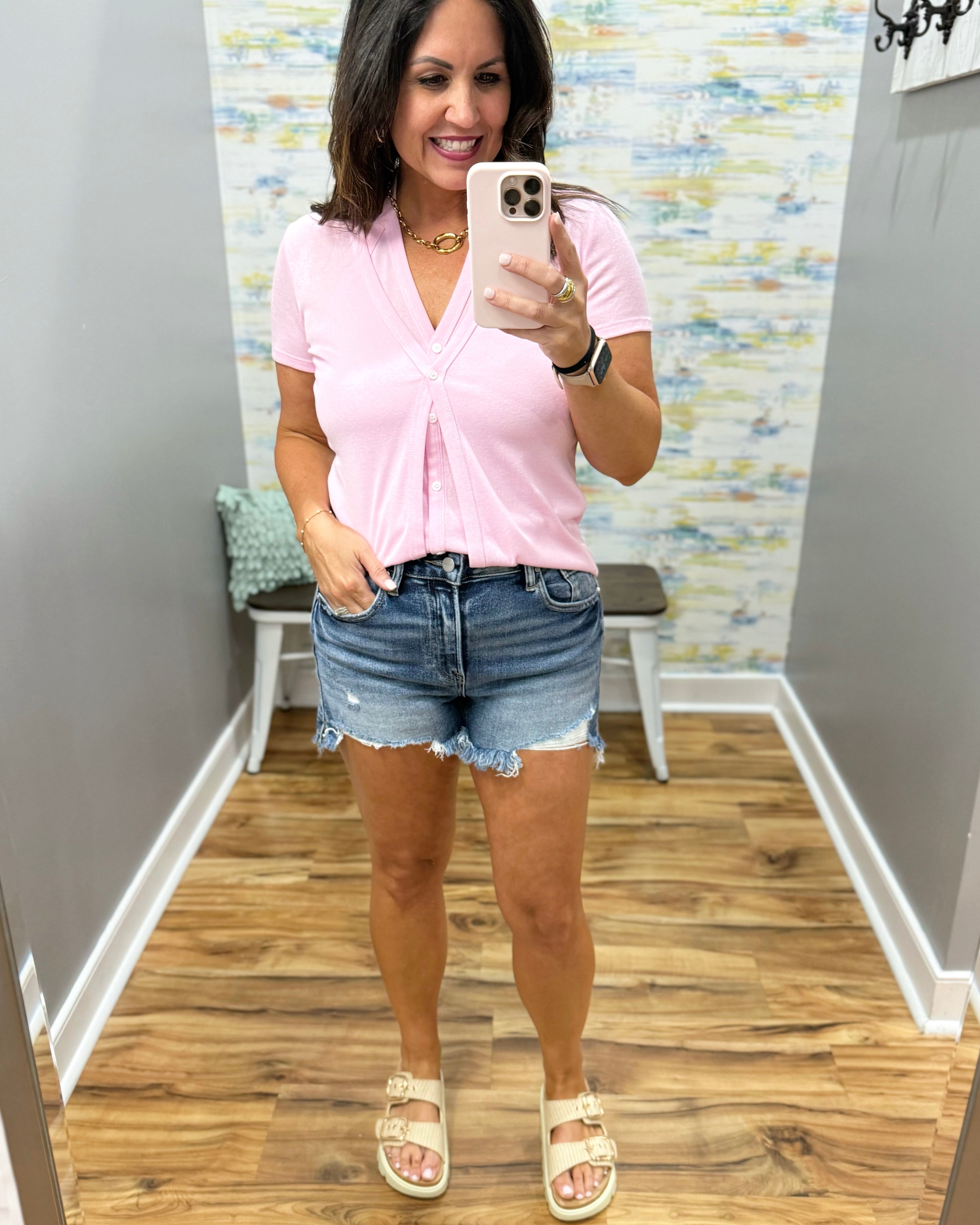 Woman taking a mirror selfie wearing a pink shirt and denim shorts in a room with a patterned wall.