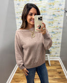 A woman takes a mirror selfie wearing a dusty rose off-the-shoulder knit sweater and blue jeans. She is holding a cream-colored smartphone in front of her face.