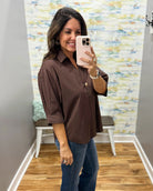 A woman takes a mirror selfie wearing a brown button-down shirt with rolled-up sleeves and blue jeans in a fitting room with a patterned wallpaper background.