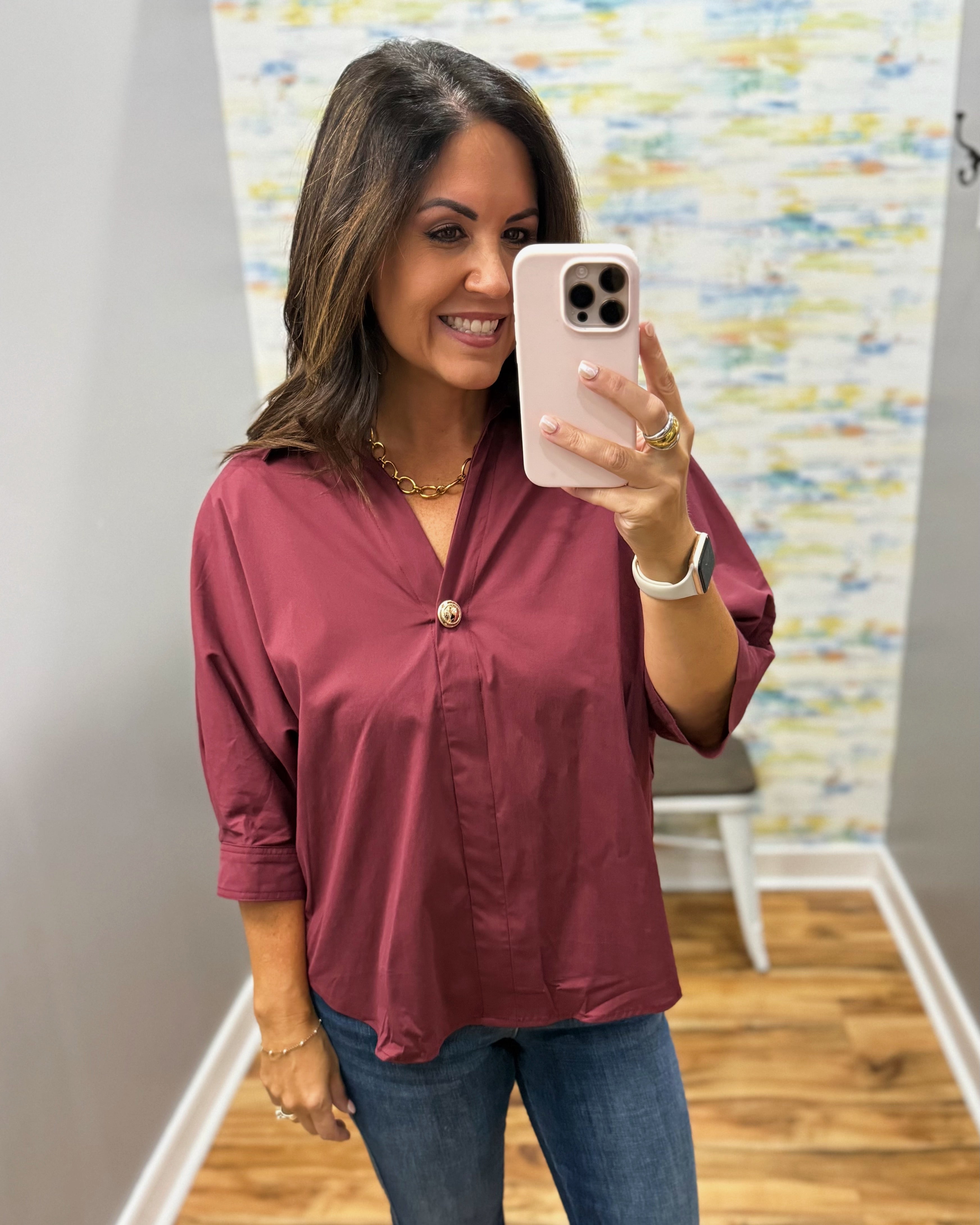 A woman takes a mirror selfie wearing a dusty rose button-down blouse with three-quarter sleeves and blue jeans in an indoor setting with a wood floor and abstract wall art.
