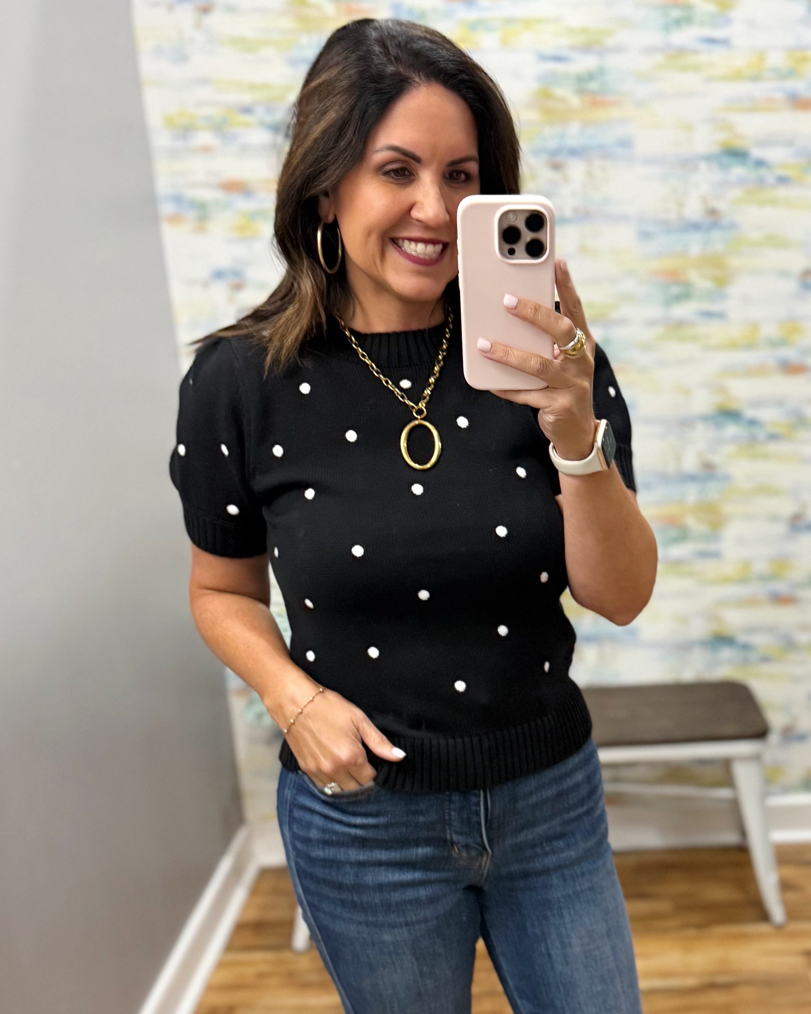 A woman takes a mirror selfie wearing a black short-sleeve knit sweater with white polka dots and blue jeans. She is wearing gold jewelry, including a long chain necklace with a large oval pendant, hoop earrings, and rings, and has a light pink smartphone in her hand.