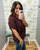 A woman taking a mirror selfie in a fitting room wearing a short-sleeve button-front top in dark burgundy and blue jeans.