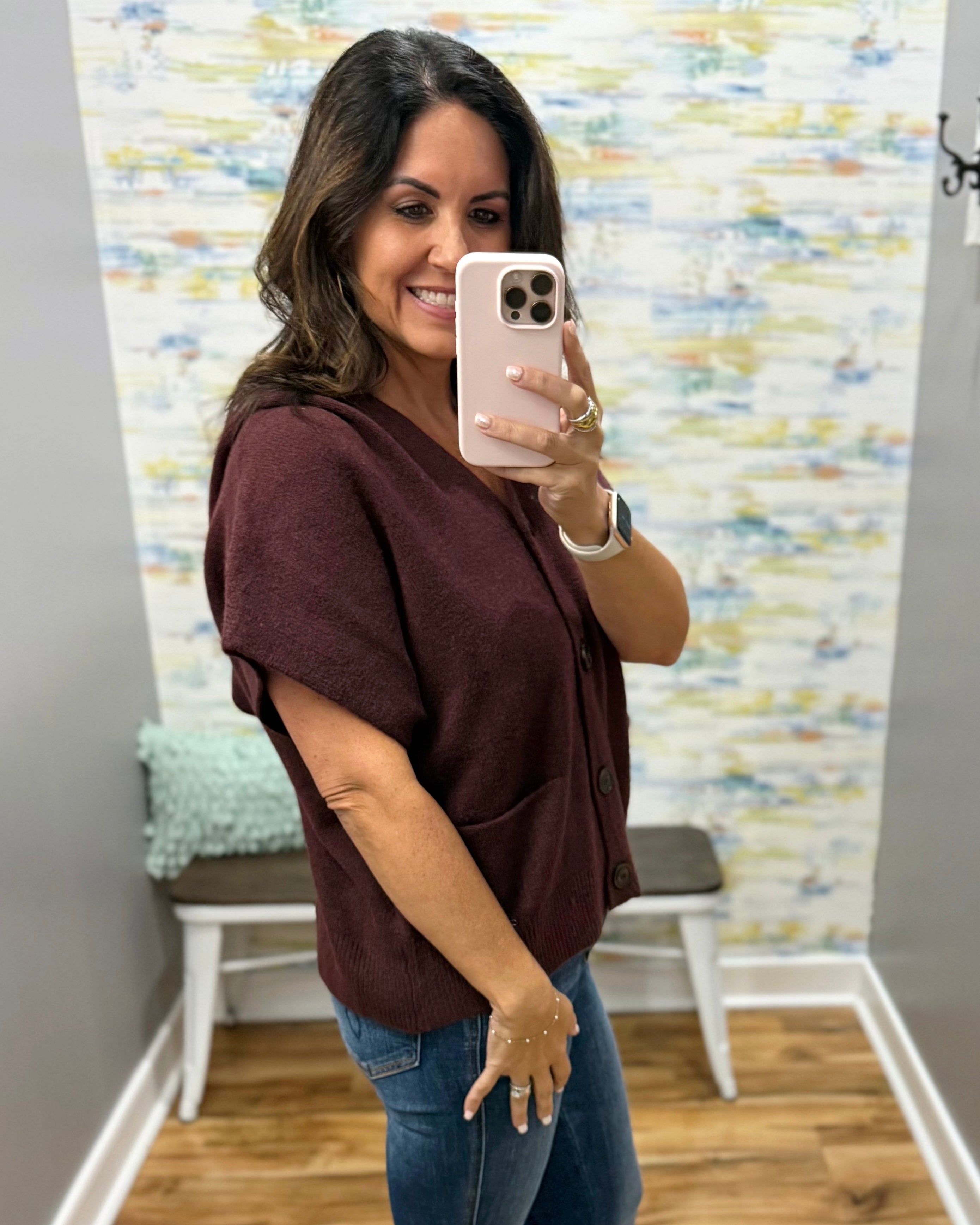 A woman taking a mirror selfie in a fitting room wearing a short-sleeve button-front top in dark burgundy and blue jeans.