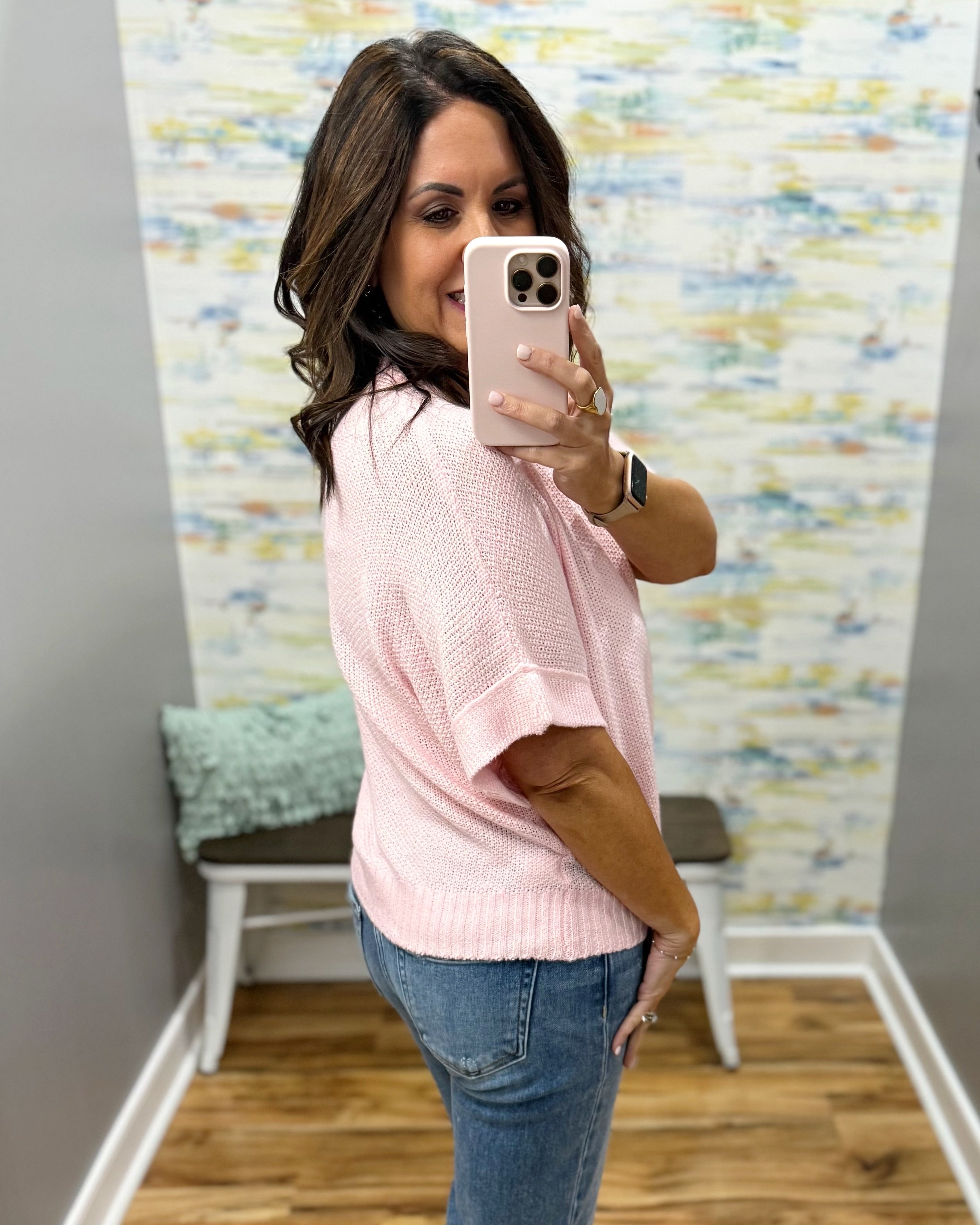 Woman taking a mirror selfie wearing a pink sweater and blue jeans in a room with a colorful wall.