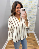 A woman takes a mirror selfie wearing a cream-colored textured button-up blouse with vertical brown stripes, paired with blue jeans.