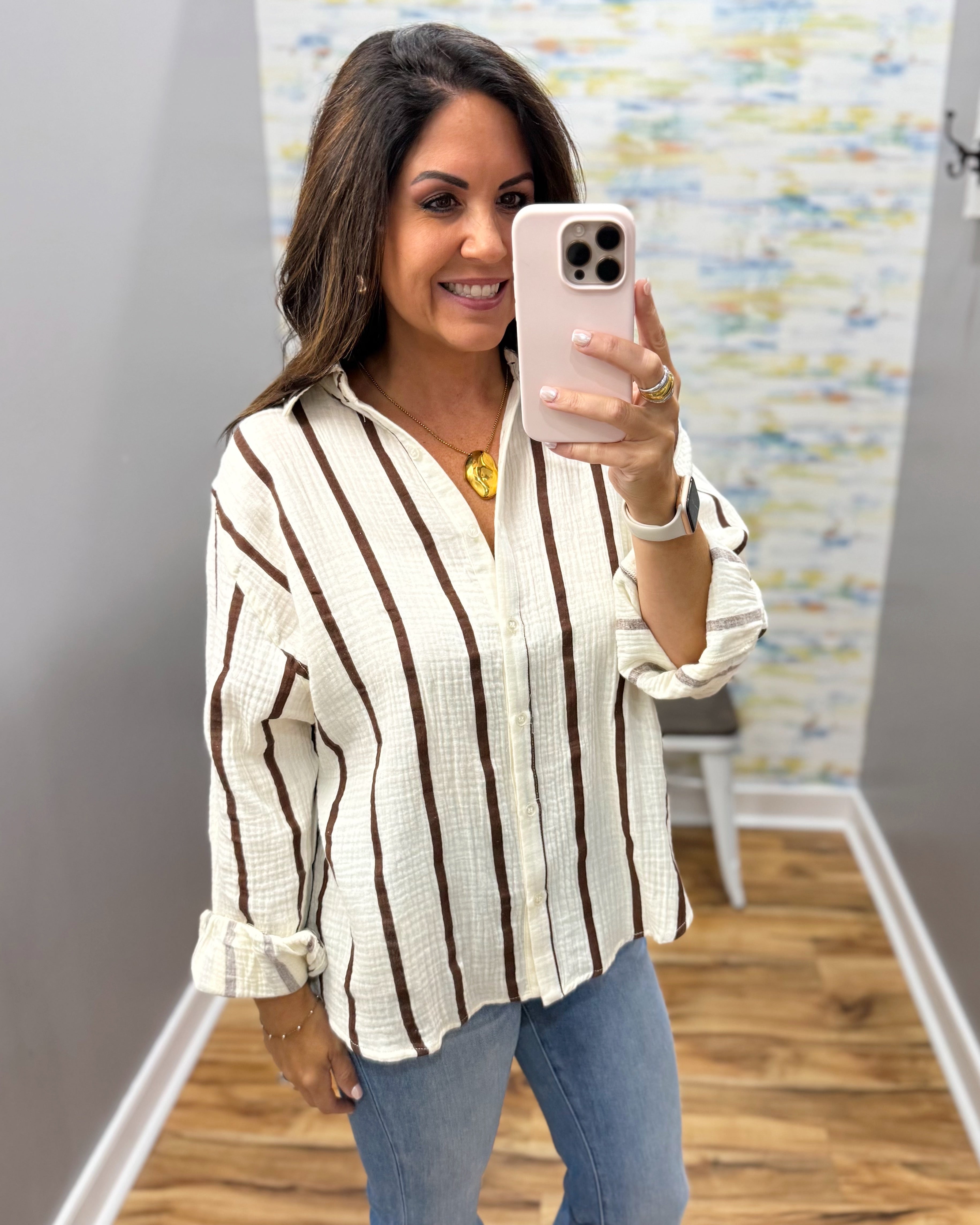 A woman takes a mirror selfie wearing a cream-colored textured button-up blouse with vertical brown stripes, paired with blue jeans.