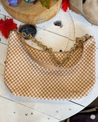 A beige and tan checkerboard shoulder bag with a gold chain strap, displayed on a rustic white wooden table.