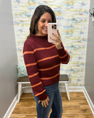 A woman taking a mirror selfie wearing a burgundy and orange striped knit sweater with blue jeans, standing in a room with a patterned wallpaper background.
