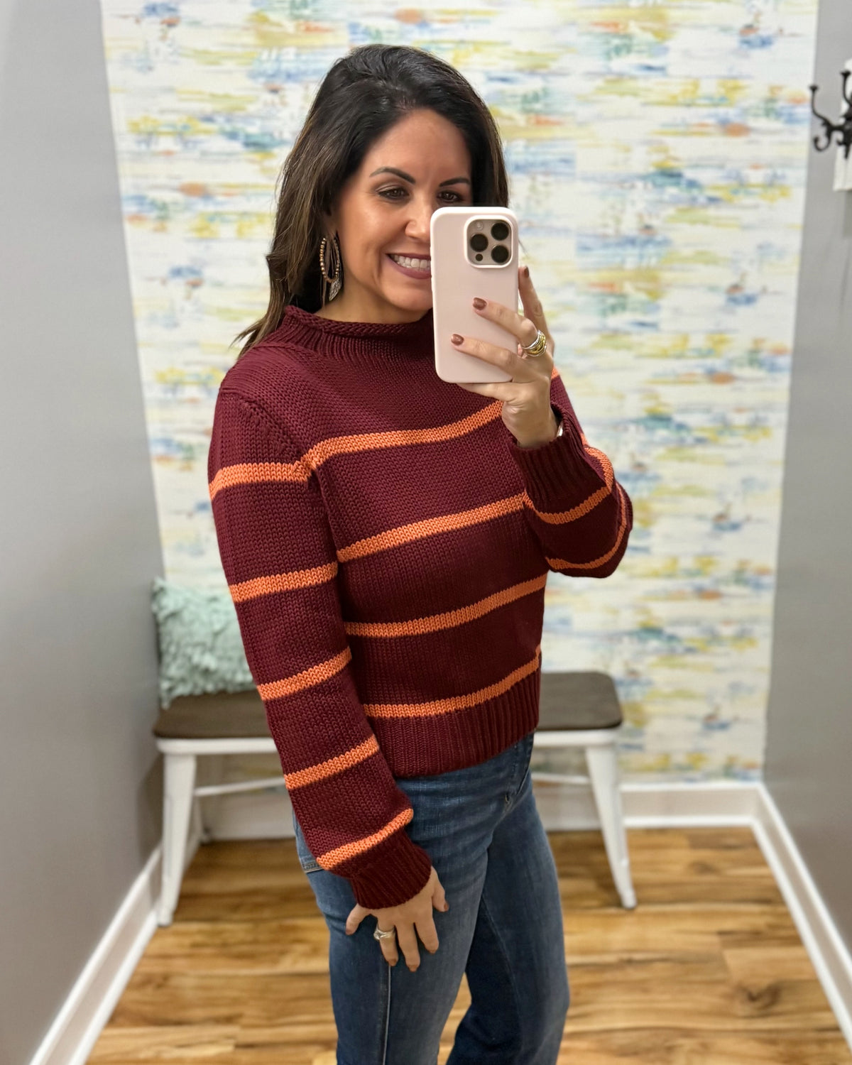 A woman taking a mirror selfie wearing a burgundy and orange striped knit sweater with blue jeans, standing in a room with a patterned wallpaper background.