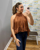 A woman takes a mirror selfie wearing a brown halter-neck ruffled top and dark blue jeans in a fitting room with a patterned wall and wooden floor.