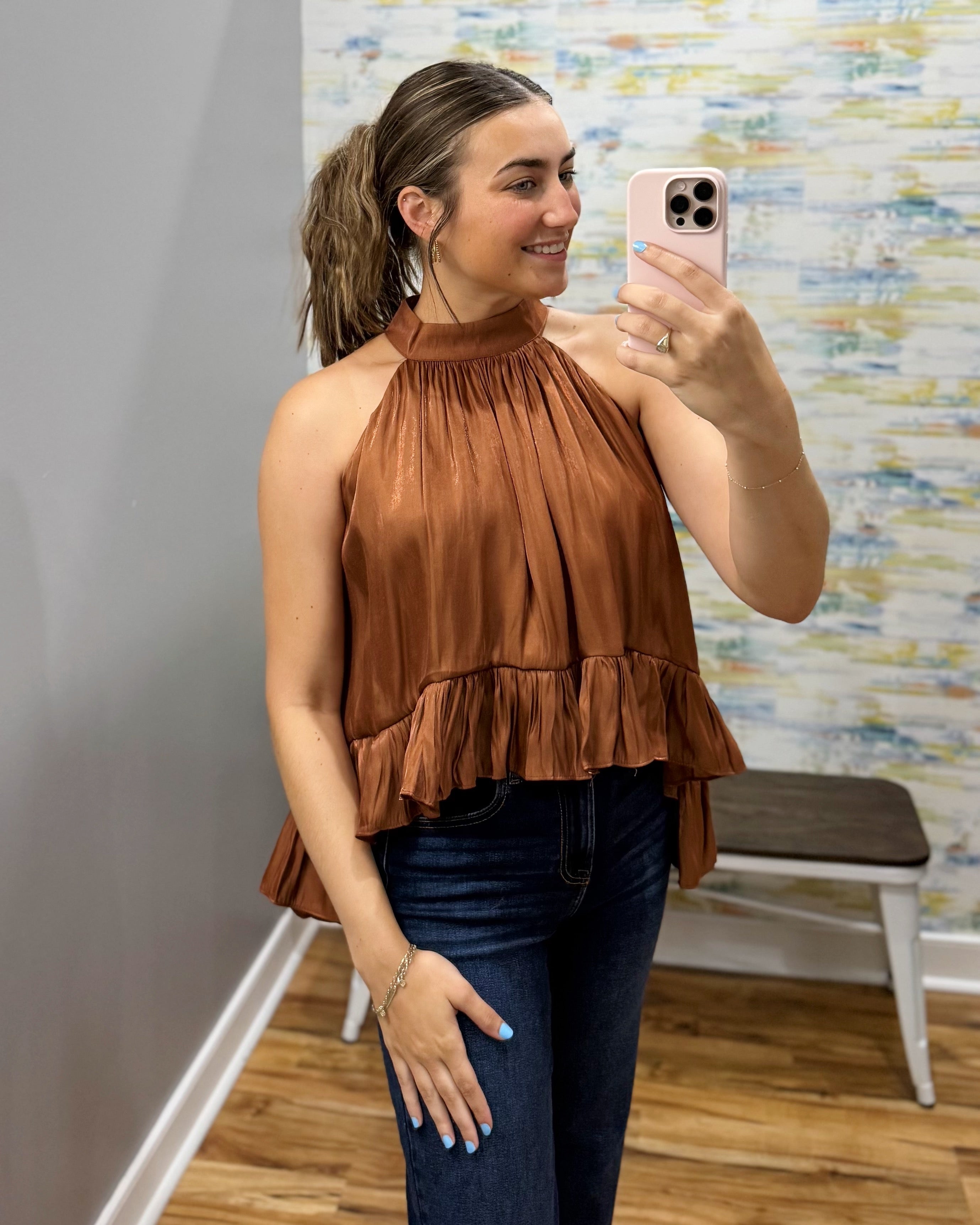 A woman takes a mirror selfie wearing a brown halter-neck ruffled top and dark blue jeans in a fitting room with a patterned wall and wooden floor.