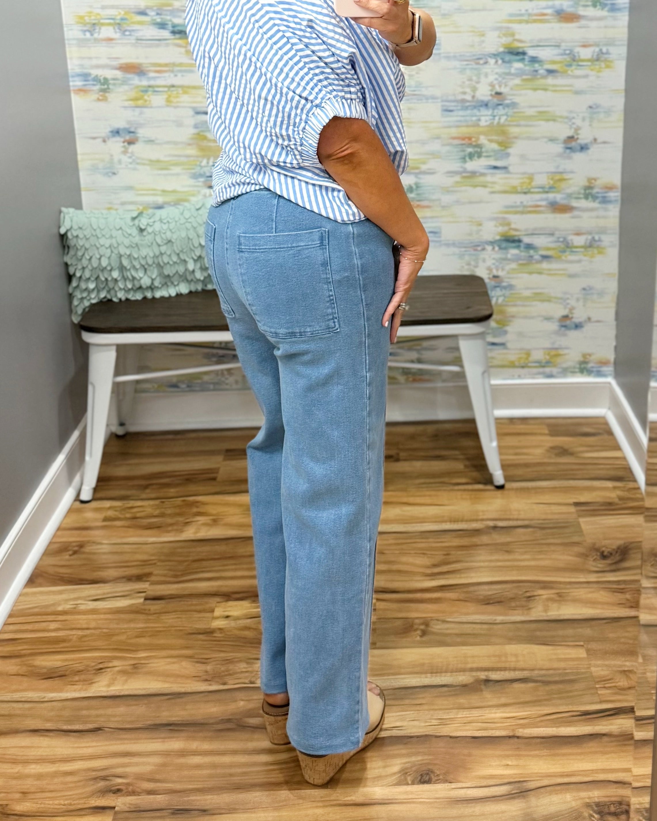 Person wearing a striped shirt and blue jeans standing in a room with wooden flooring and a colorful wall.