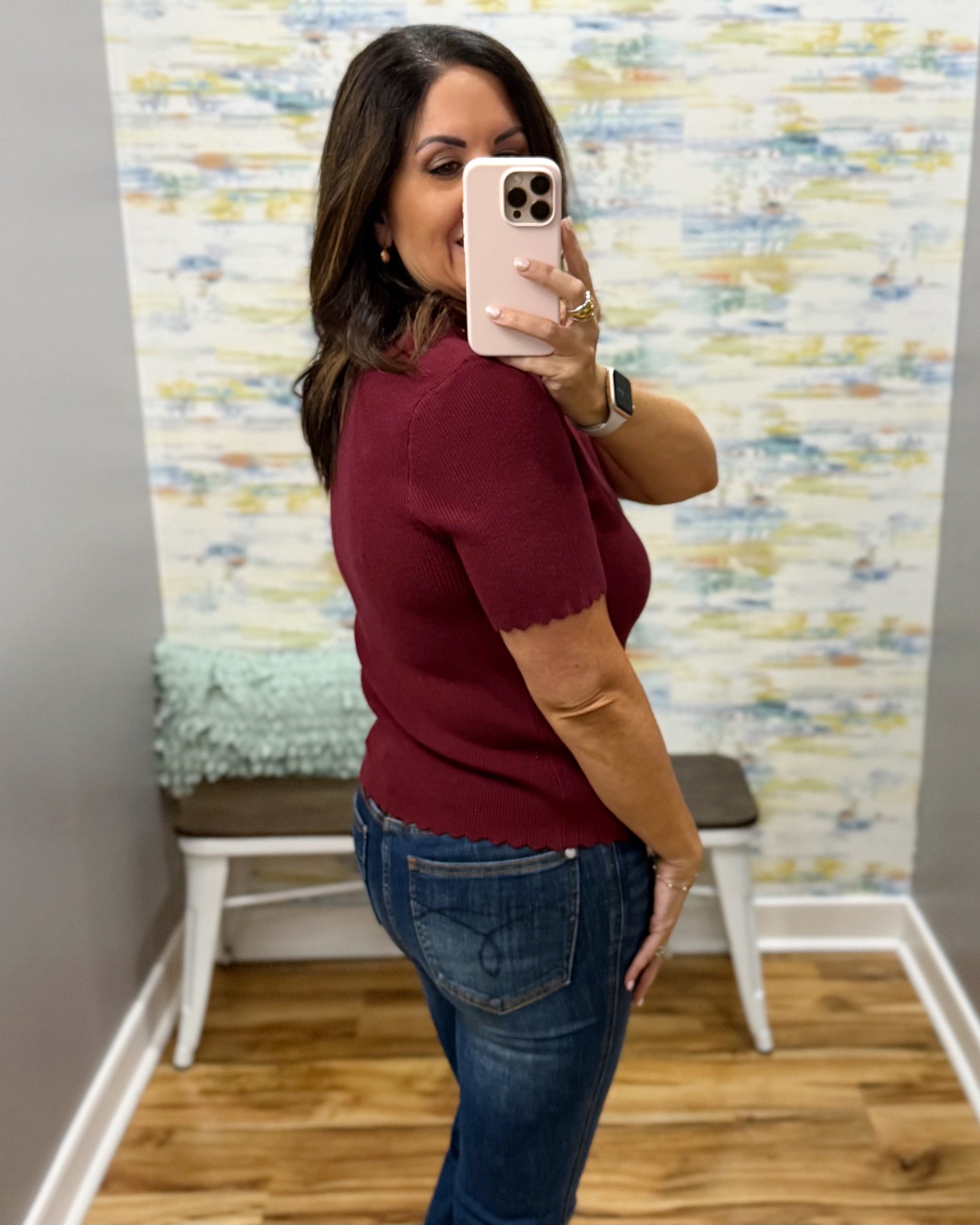 A woman takes a mirror selfie in a fitting room wearing a short-sleeve burgundy knit top and blue jeans.
